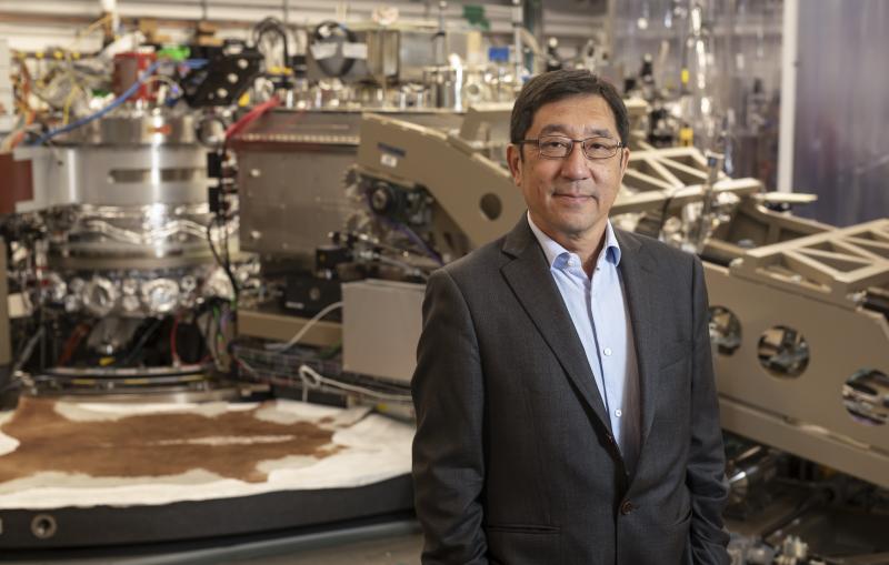 A man in a blue shirt and gray suit poses in front of a large scientific apparatus.